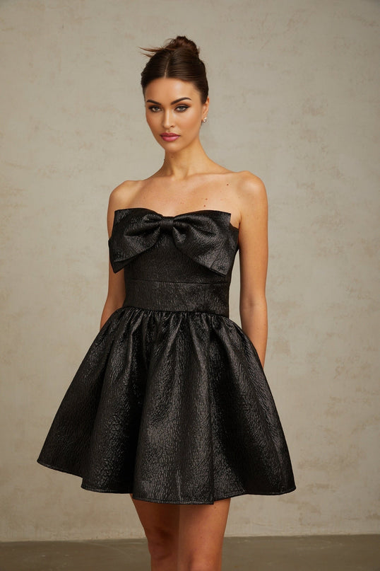 Woman wearing a black mini dress with metallic sheen and large bow detail