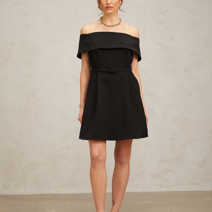 Woman wearing a black offshoulder mini dress with a belt cinching the waist standing against a plain background