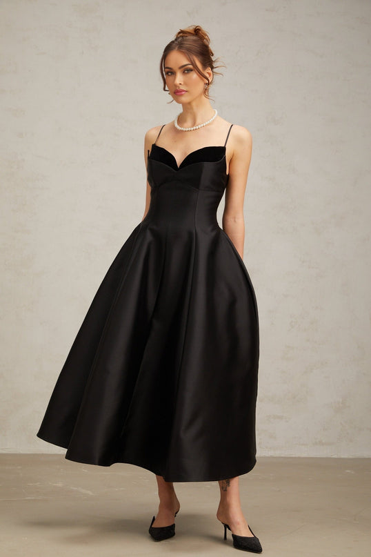 Woman wearing a black pleated midi dress with flared skirt and fitted bodice standing against plain background