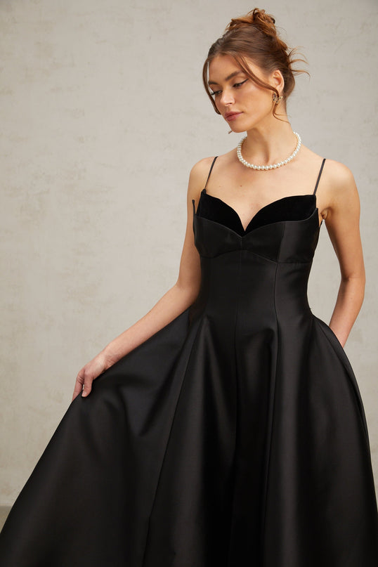 Woman wearing a black pleated midi dress with flared skirt and fitted bodice standing in a neutral background