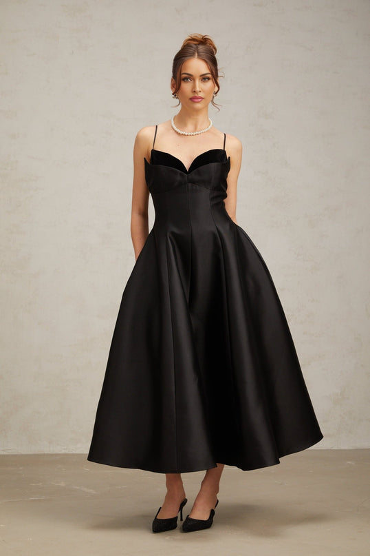 Woman wearing a black pleated midi dress with flared skirt and sleeveless design standing against plain background