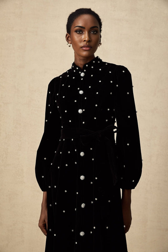 Woman wearing a black velvet midi dress with embellishments standing against a plain background
