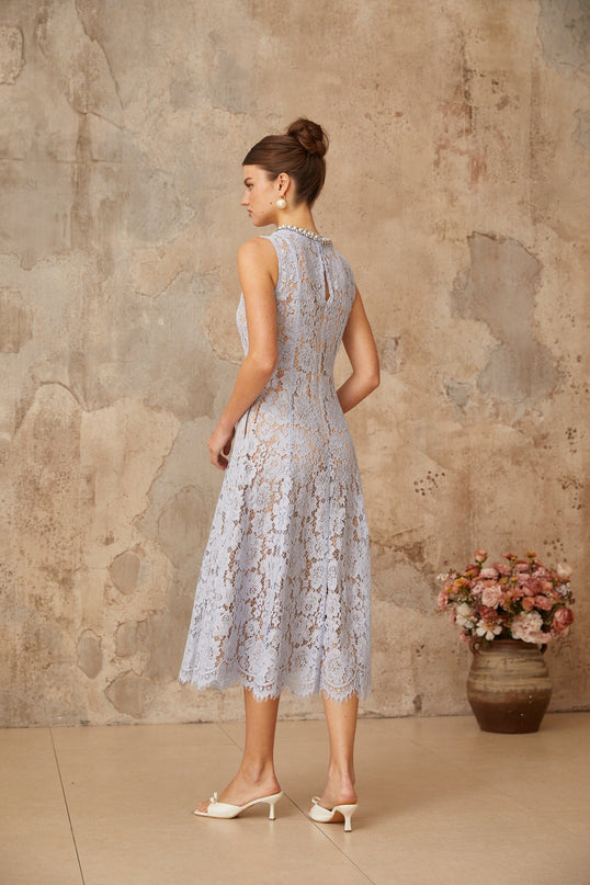 Woman wearing a blue midi dress with floral lace pattern and embellishments standing in front of a plain background