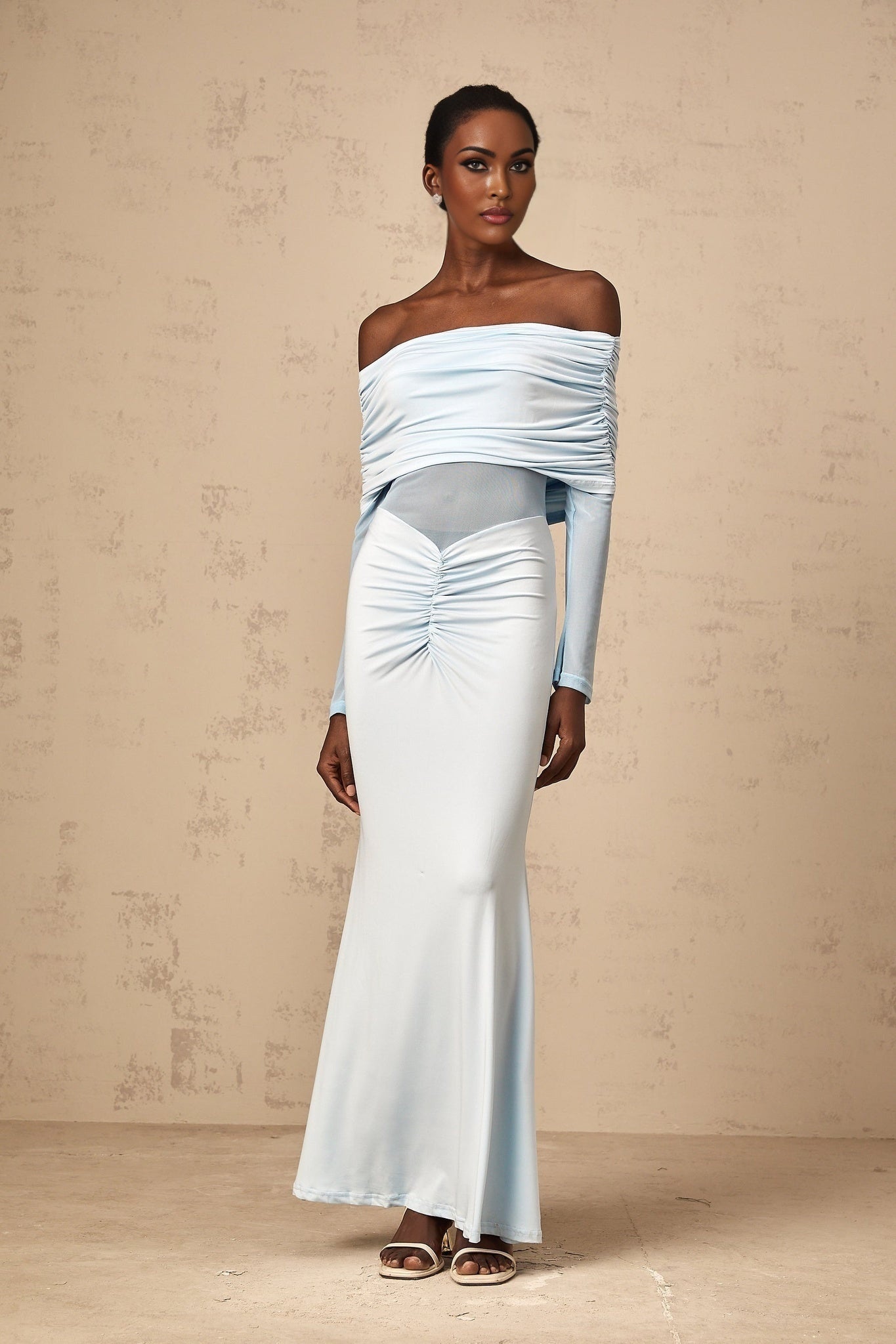 Woman wearing a blue offshoulder maxi dress with ruched detailing standing against a plain background