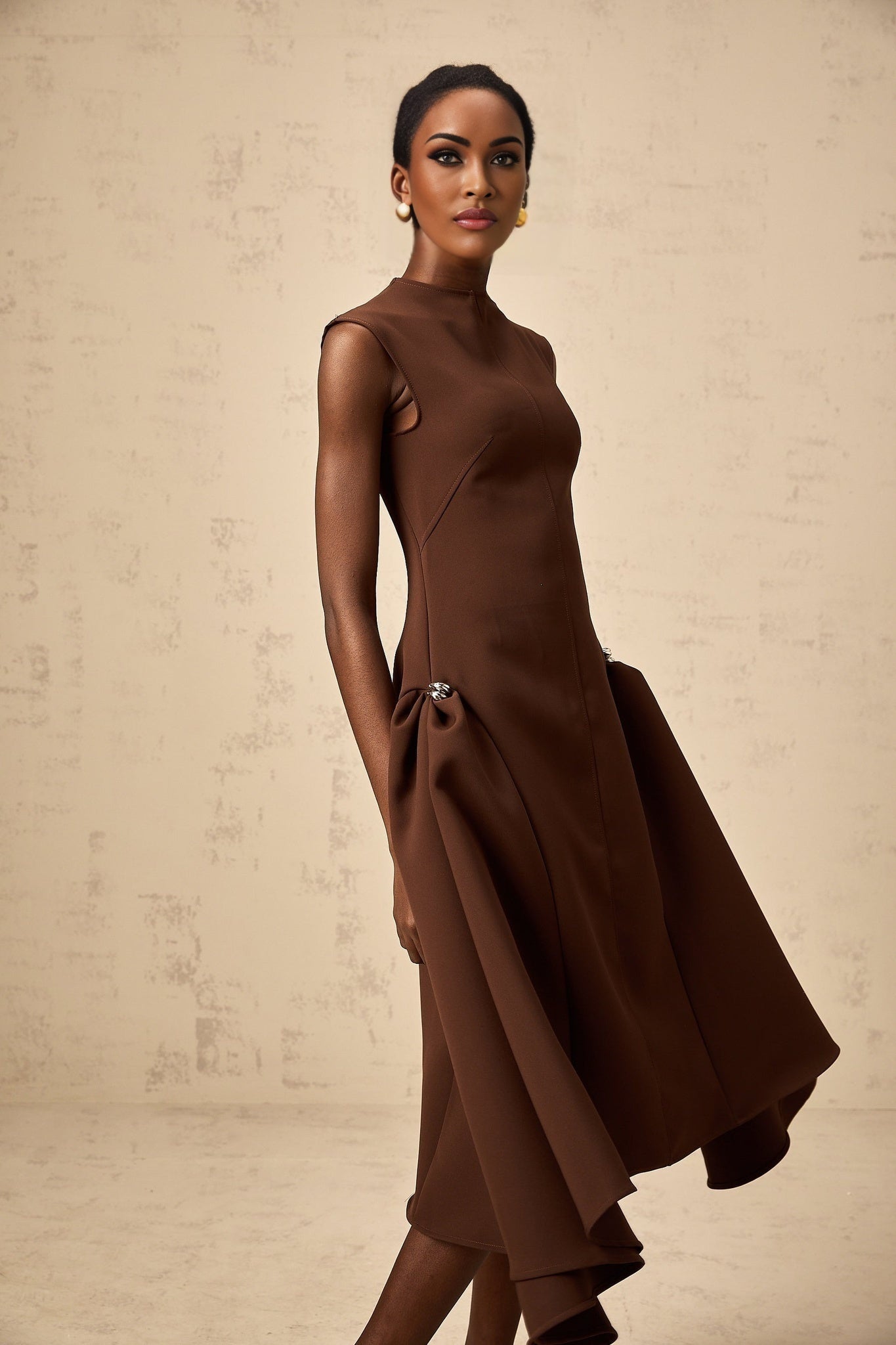 Woman wearing a brown asymmetrical midi dress with a flowy skirt and fitted bodice standing against a plain background
