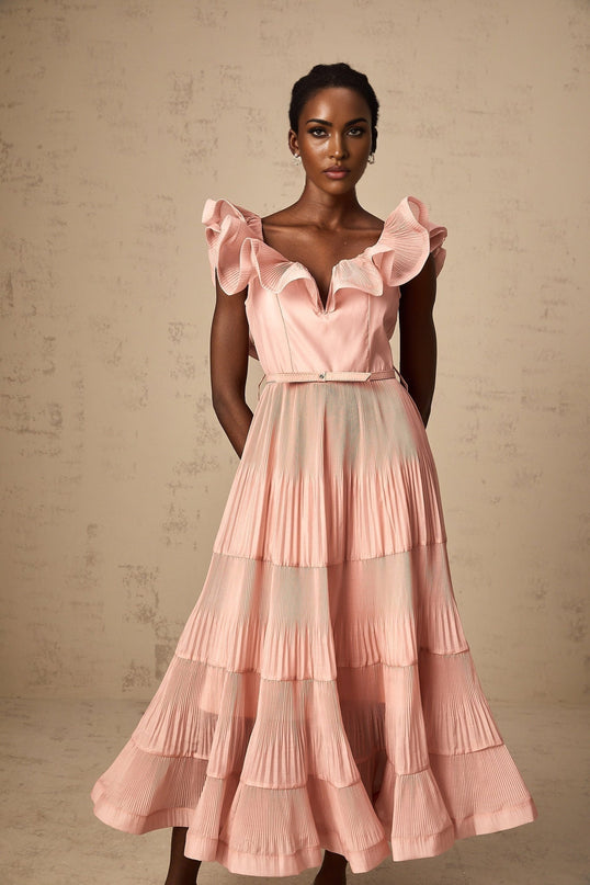 Woman wearing a coralpink midi dress with pleated details and frilled accents standing against a plain background