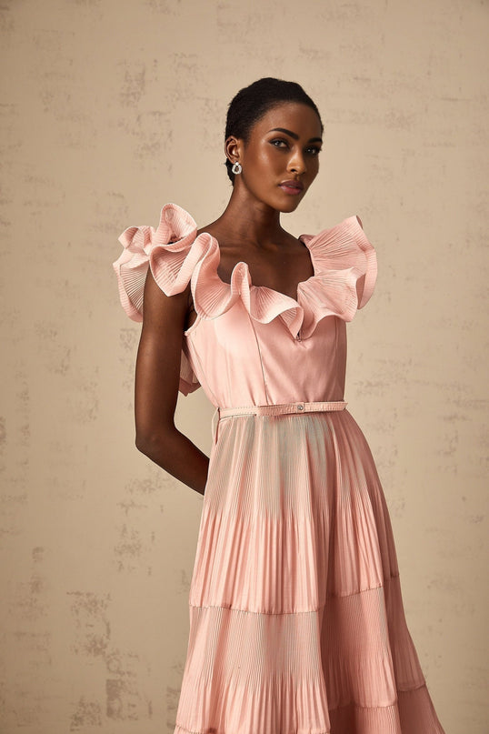Woman wearing a coralpink midi dress with pleated fabric and frilly details standing in a neutral background