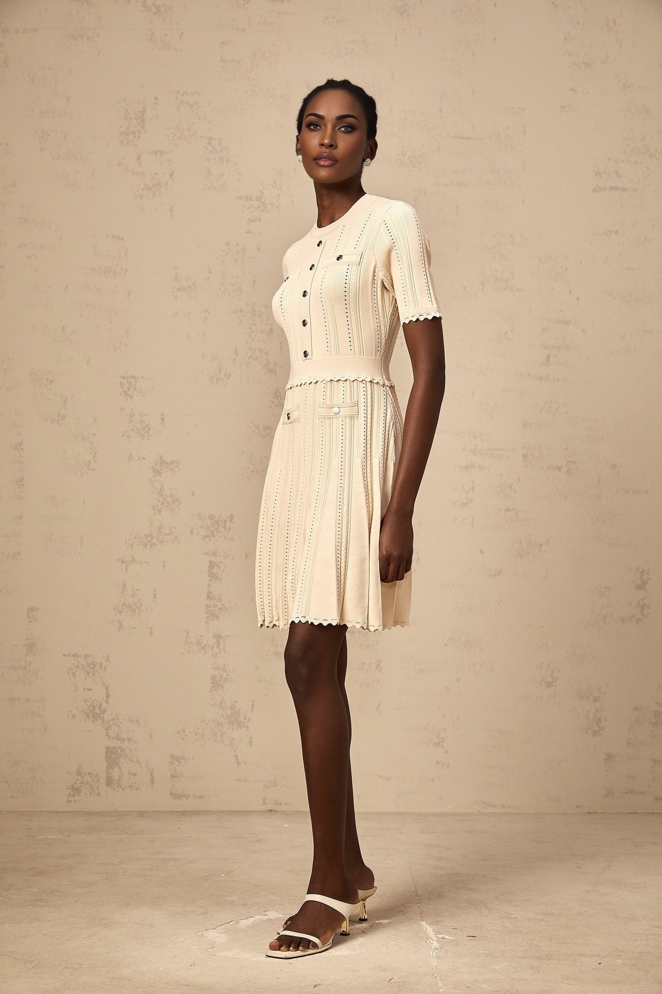 Woman wearing a lightcolored knit mini dress with pleated skirt and delicate pointelle trim detail standing in a neutral setting
