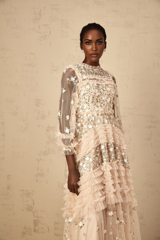 Woman wearing a long floral embroidered tulle maxi dress with tiered skirt in soft pastel colors against a plain background