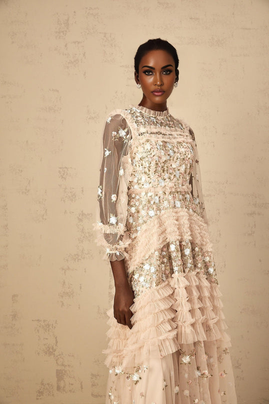 Woman wearing a long floral embroidered tulle maxi dress with tiered skirt in soft pastel colors against a plain background