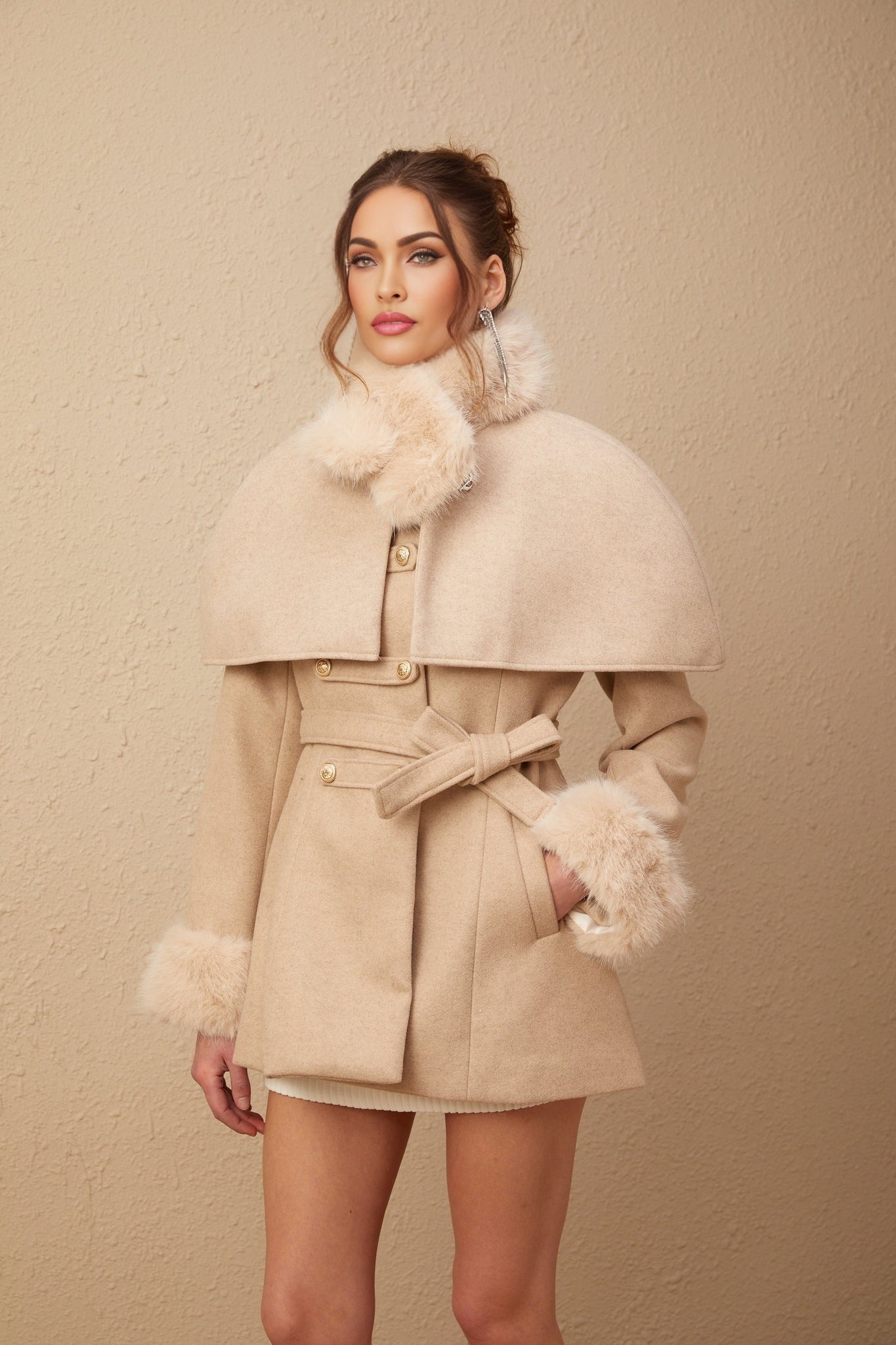 Woman wearing a long white faux fur coat with a detachable cape collar standing in a studio setting