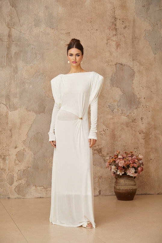 Woman wearing a long white maxi dress with draped and shirred details standing against a plain background