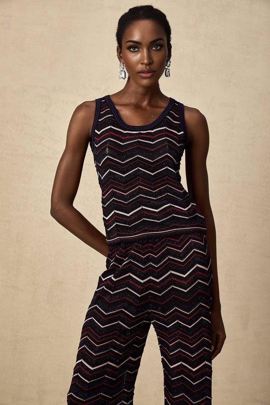 Woman wearing a matching set of zigzag patterned crochet knit top and trousers in beige color standing against a plain background