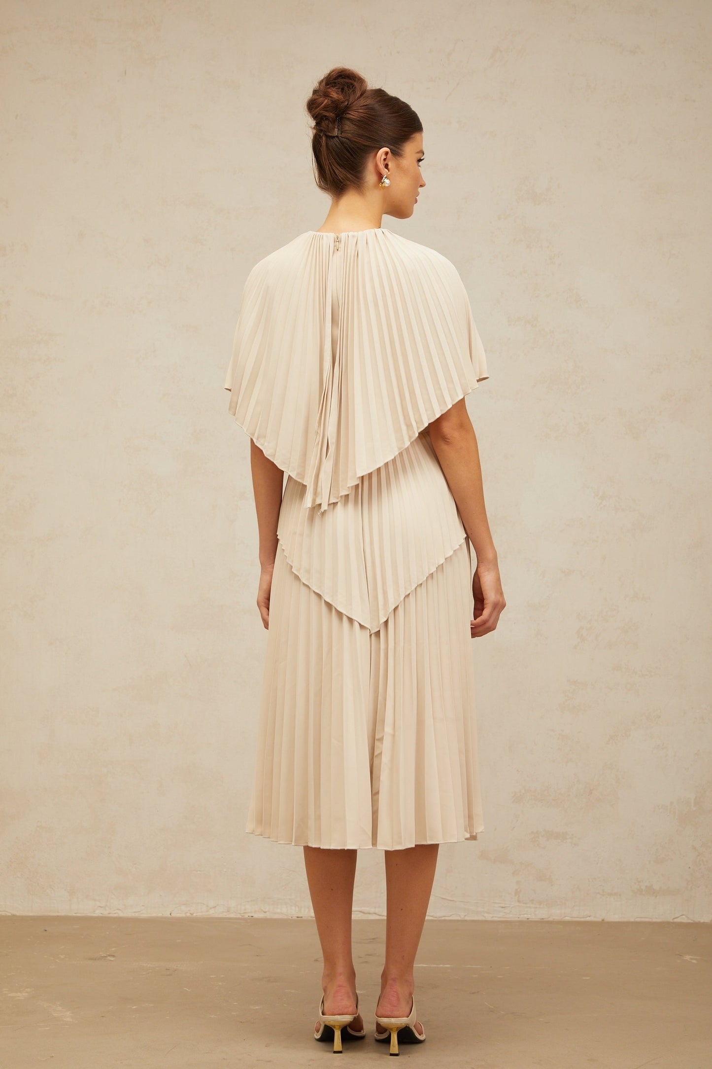 Woman wearing a neutral colored cape style pleated midi dress with flowing fabric and elegant silhouette