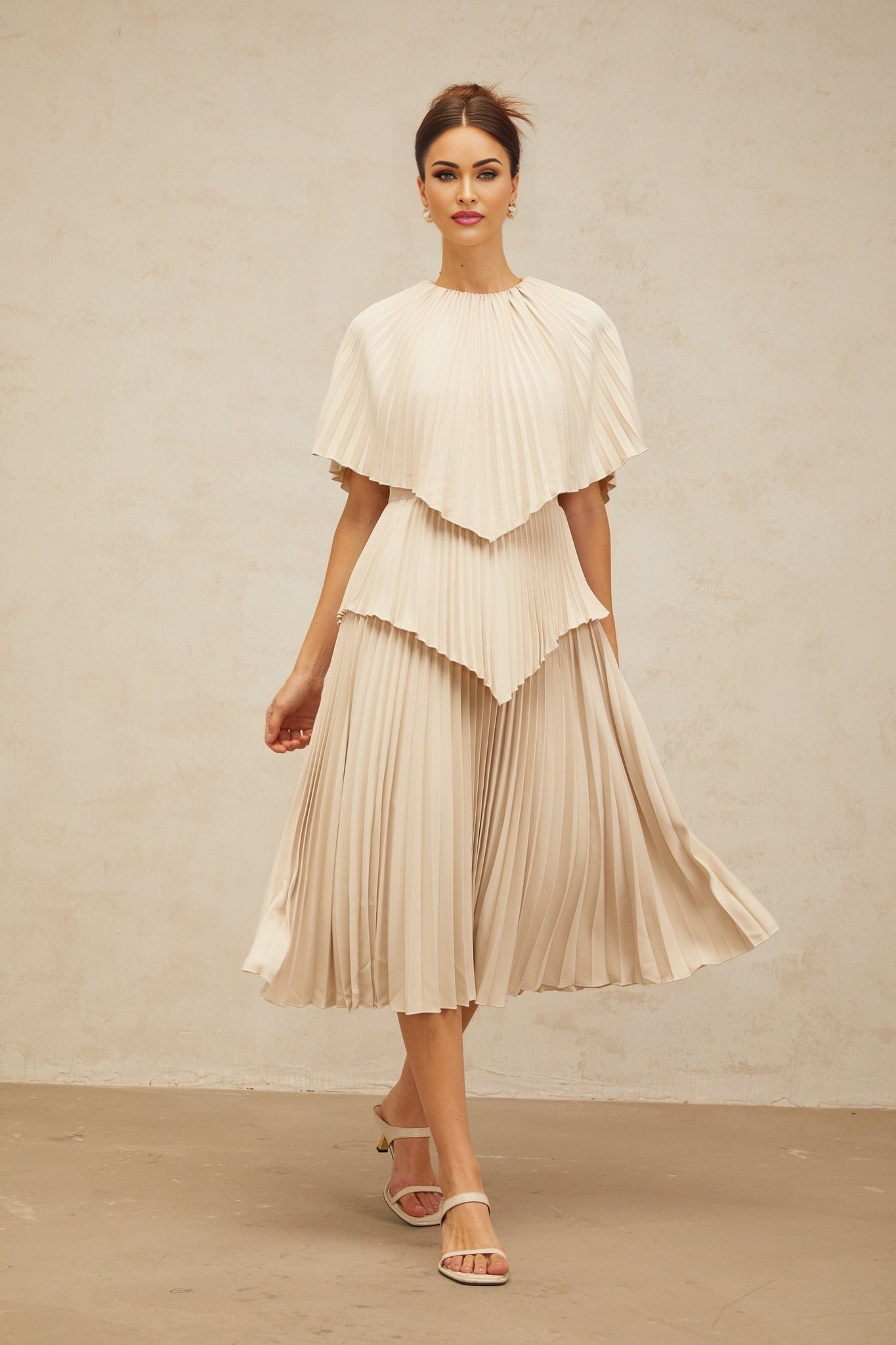 Woman wearing a neutral colored cape style pleated midi dress with flowy silhouette and elegant draping