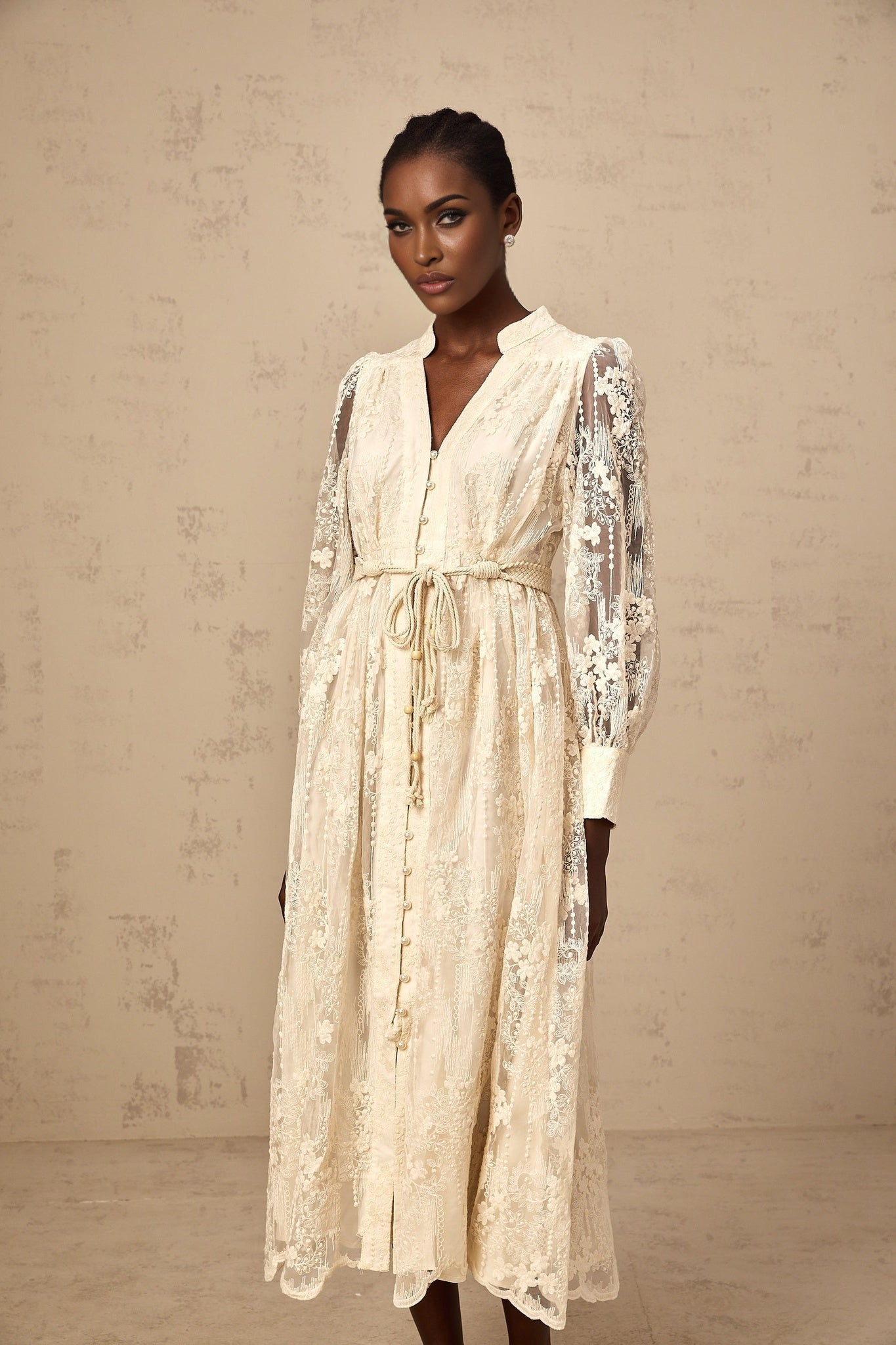 Woman wearing a neutralcolored midi dress with lace embroidery details standing against a plain background