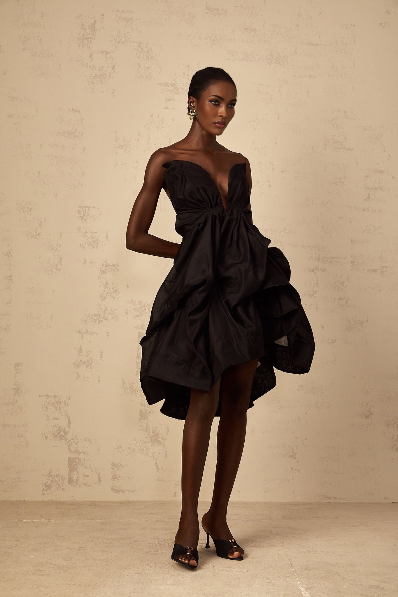 Woman wearing a short asymmetrical pleated cocktail dress with one shoulder strap in a dark color against a plain background