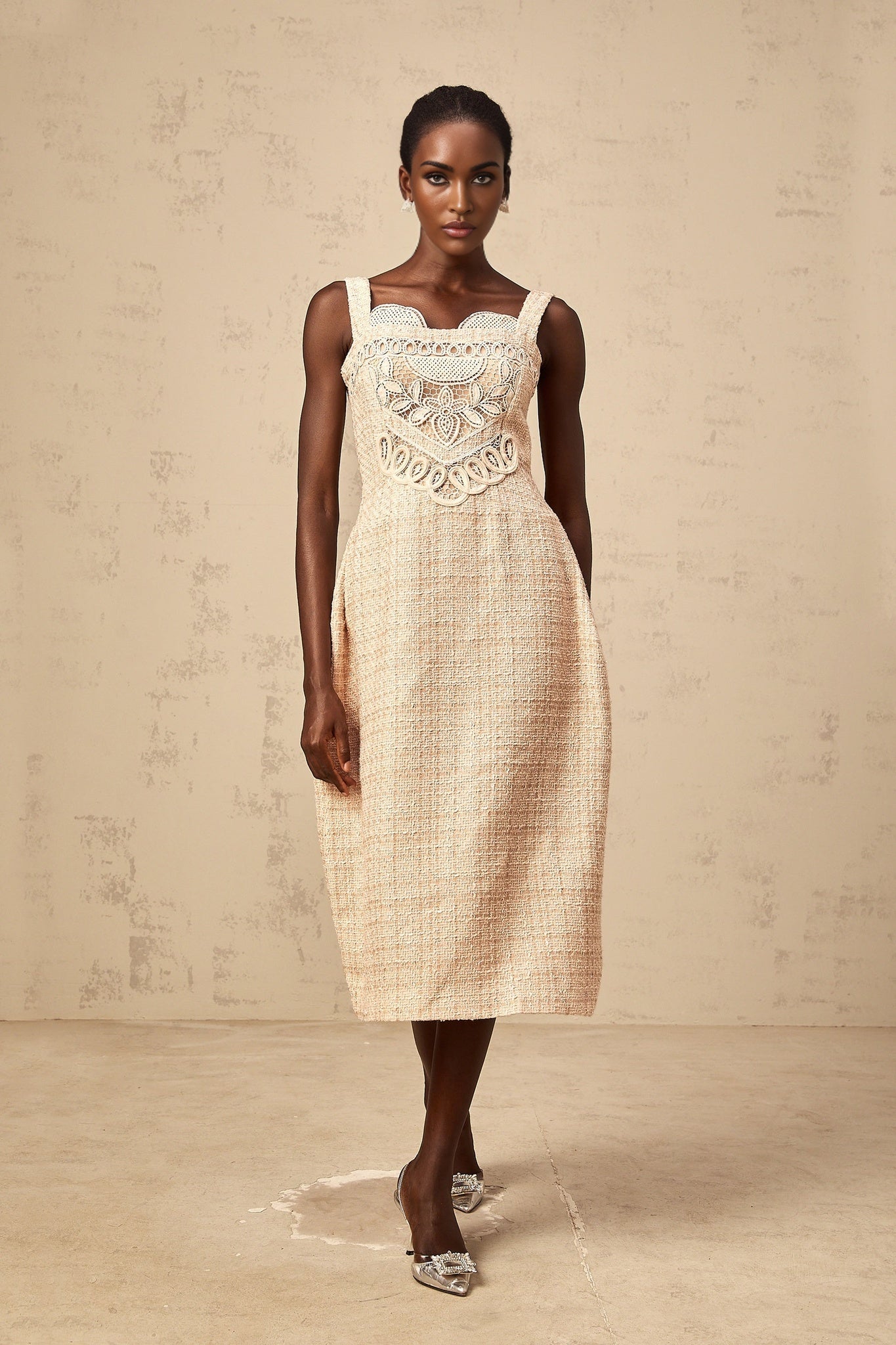 Woman wearing a short beige dress with intricate embroidery details and woven patterns standing against a plain background