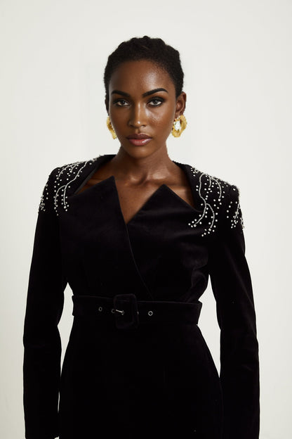 Woman wearing a short black velvet dress with crystal embellishments and thin straps standing in front of plain background