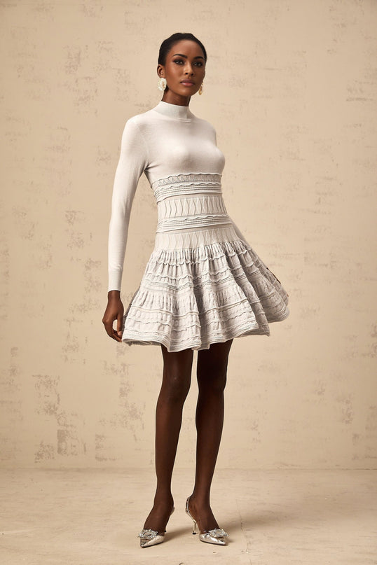 Woman wearing a short grey dress with ruffled details and ribbed texture standing against plain background