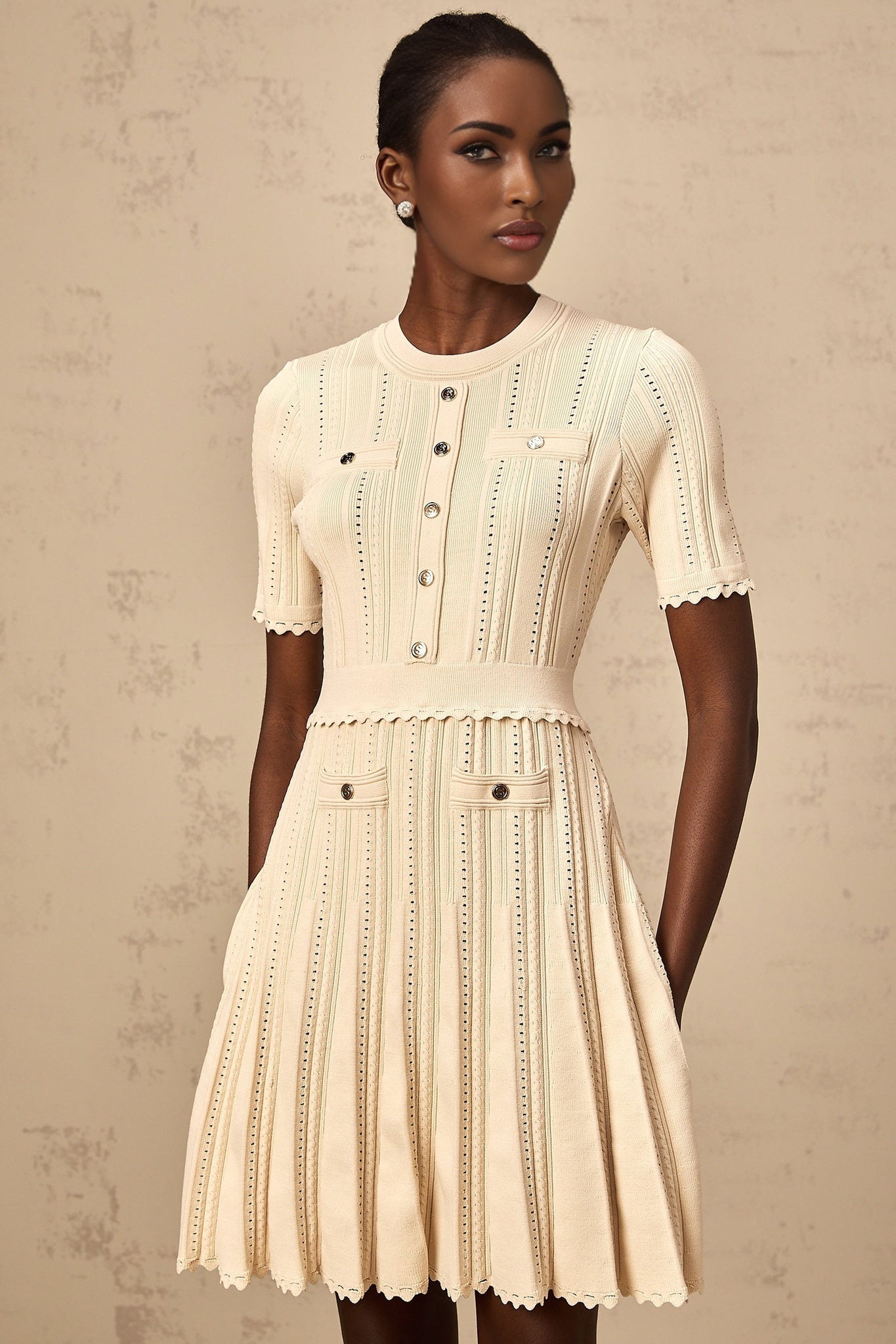 Woman wearing a short neutral colored knit dress with pleated skirt and delicate pointelle trim detail