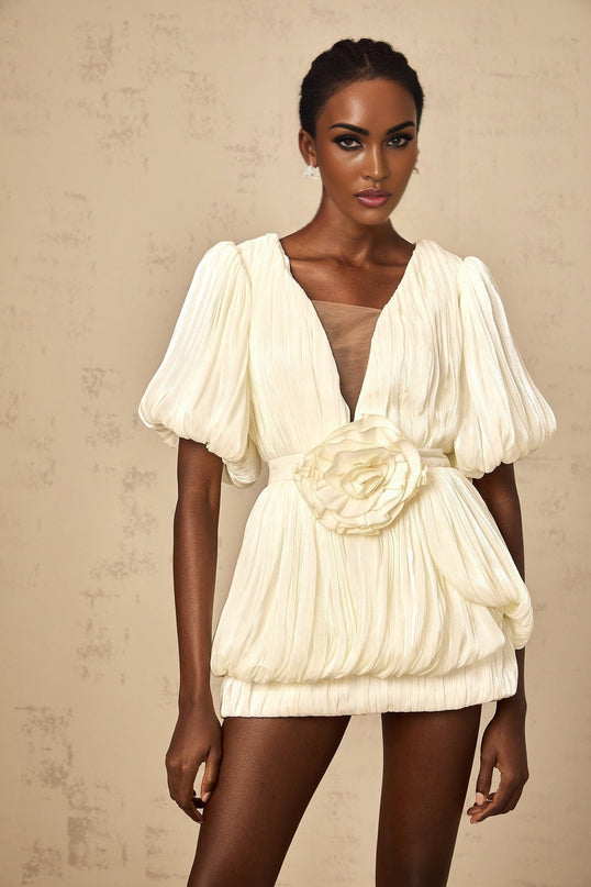 Woman wearing a short neutralcolored pleated dress with floral appliques standing against a plain background