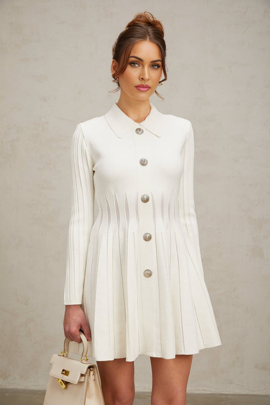 Woman wearing a short white pleated knit dress with long sleeves and a high neckline standing against a plain background