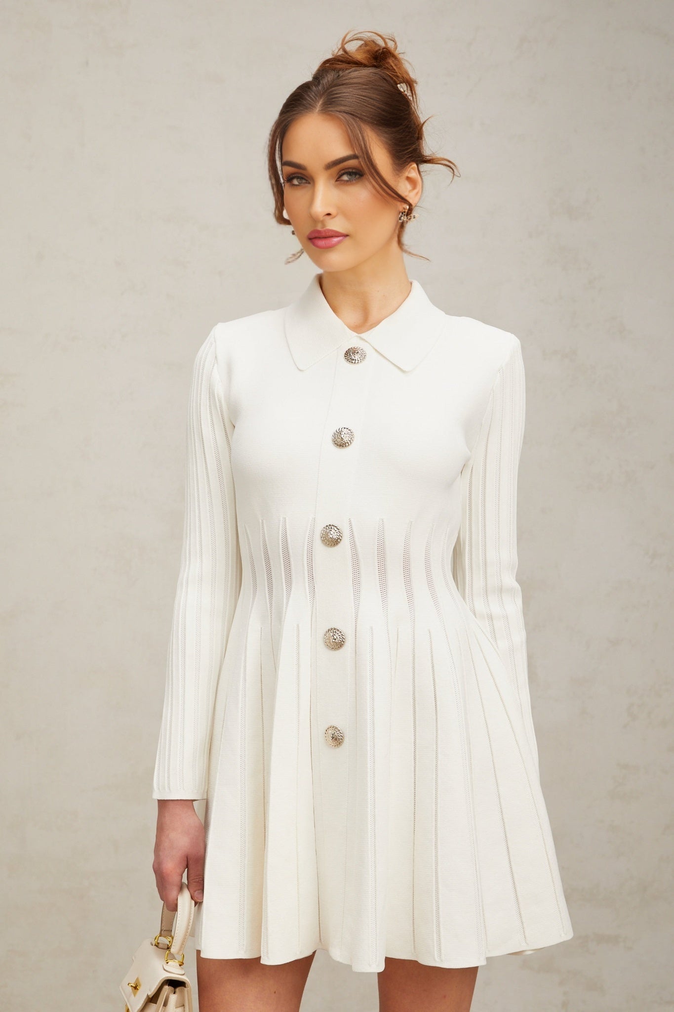 Woman wearing a short white pleated knit dress with long sleeves standing against a plain background
