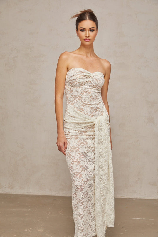 Woman wearing a white lace midi dress with draped details and delicate floral patterns standing against a plain background