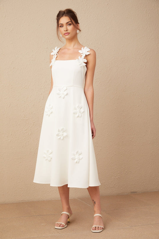 Woman wearing a white midi dress with square neckline and faux flower details standing against plain background
