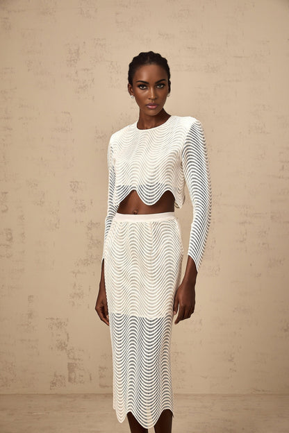 Woman wearing a white twopiece outfit consisting of a wavy woven top and matching skirt standing against a plain background
