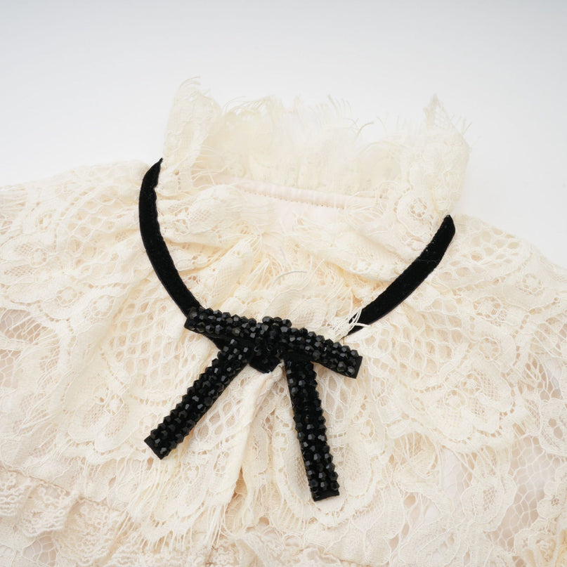Woman wearing an offwhite blouse with floral lace pattern and large bow detail at neckline