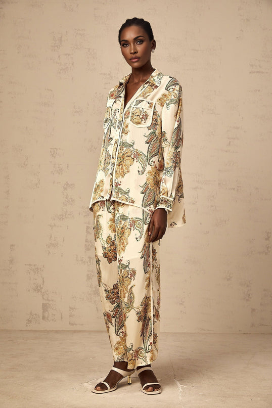 Woman wearing beige floral print shirt and matching trousers with neutral tones standing against plain background