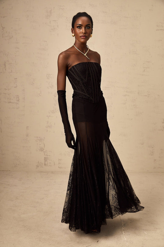 Woman wearing black floral lace corset matching skirt and gloves set against plain background