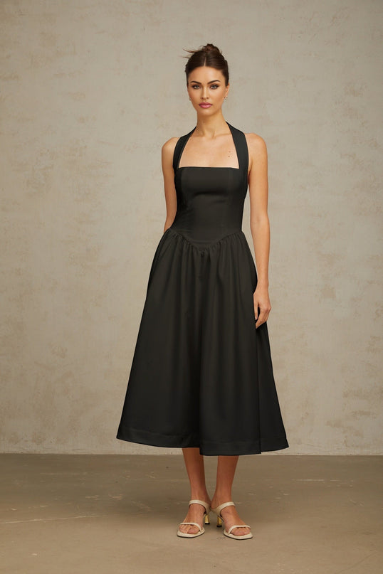 Woman wearing black halterneck pleated midi dress with fitted waist and flowing skirt