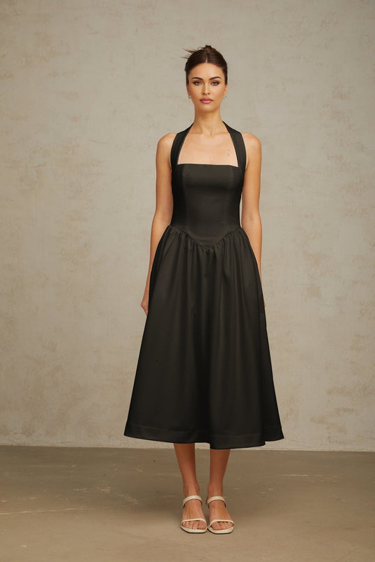 Woman wearing black halterneck pleated midi dress with fitted waist and flowy skirt