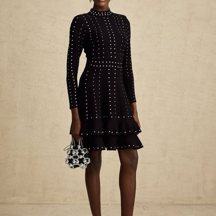 Woman wearing black knitted mini dress with ruffled details and embellishments standing against plain background