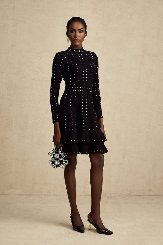 Woman wearing black knitted mini dress with ruffled details and embellishments standing against plain background