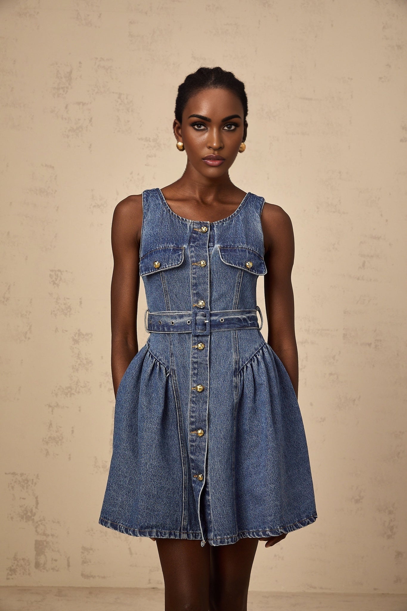 Woman wearing blue denim mini dress with belt cinched at waist standing against plain background