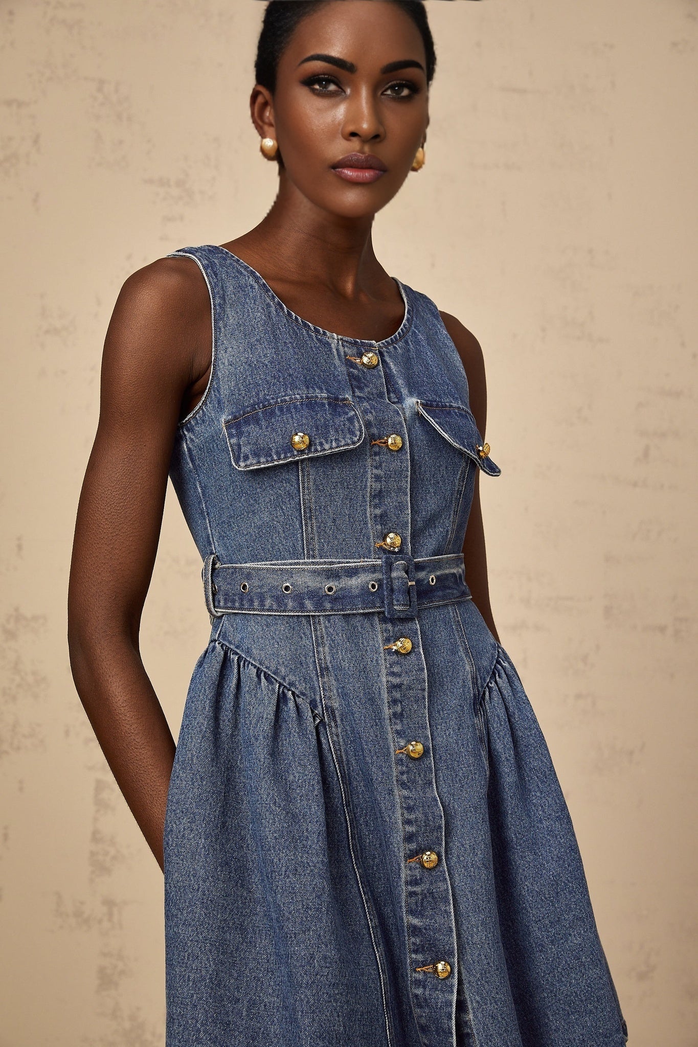 Woman wearing blue denim mini dress with belt cinched at waist standing against plain background