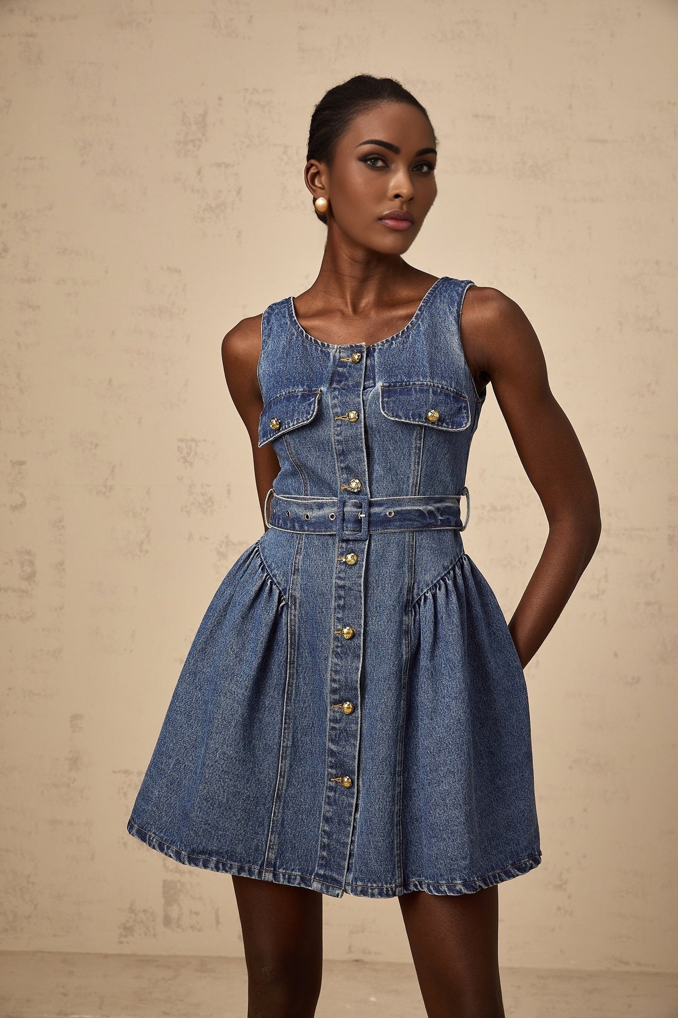 Woman wearing blue denim mini dress with belt cinched at waist standing against plain background
