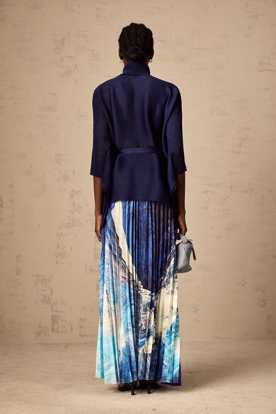 Woman wearing blue patterned cape and matching skirt set with creased texture standing against plain background