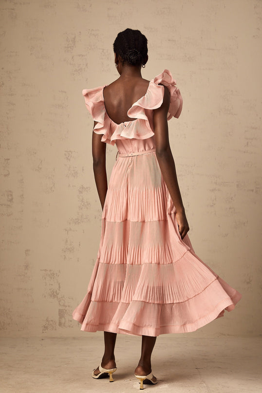 Woman wearing coralpink pleated midi dress with frilled details and sleeves standing against plain background