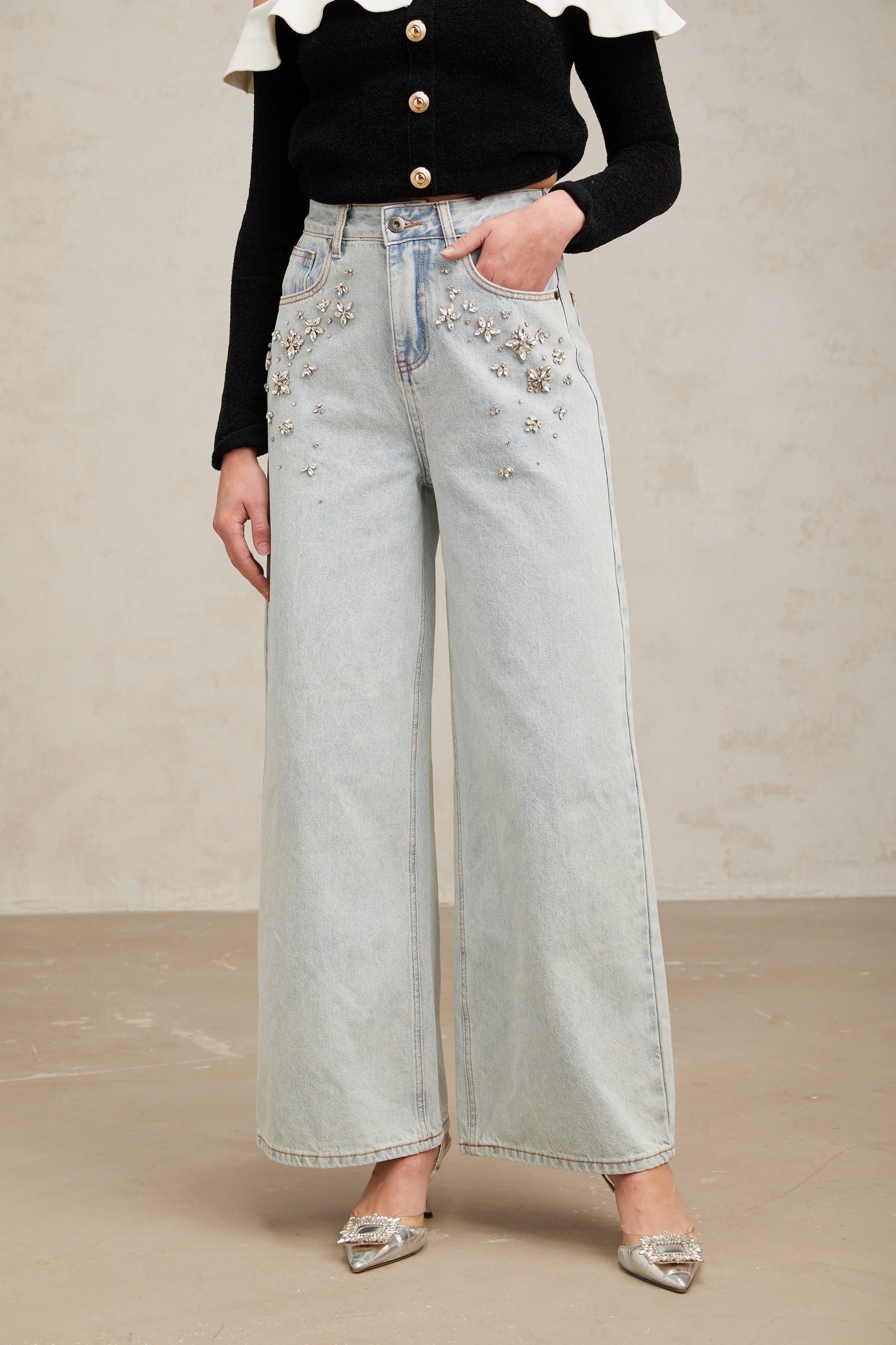 Woman wearing light blue jeans with decorative embellishments on the legs standing against a plain background