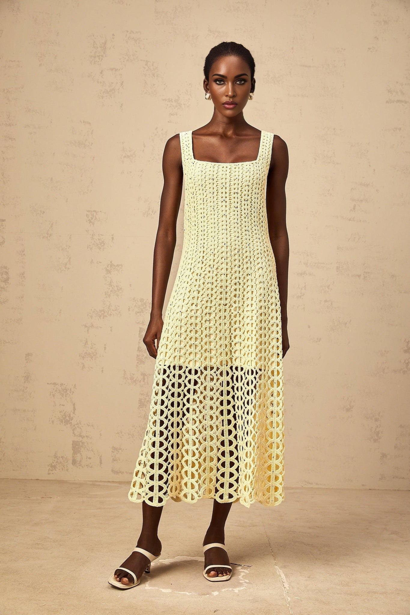 Woman wearing light yellow crochet knit midi dress with cutout details and embellishments standing against plain background