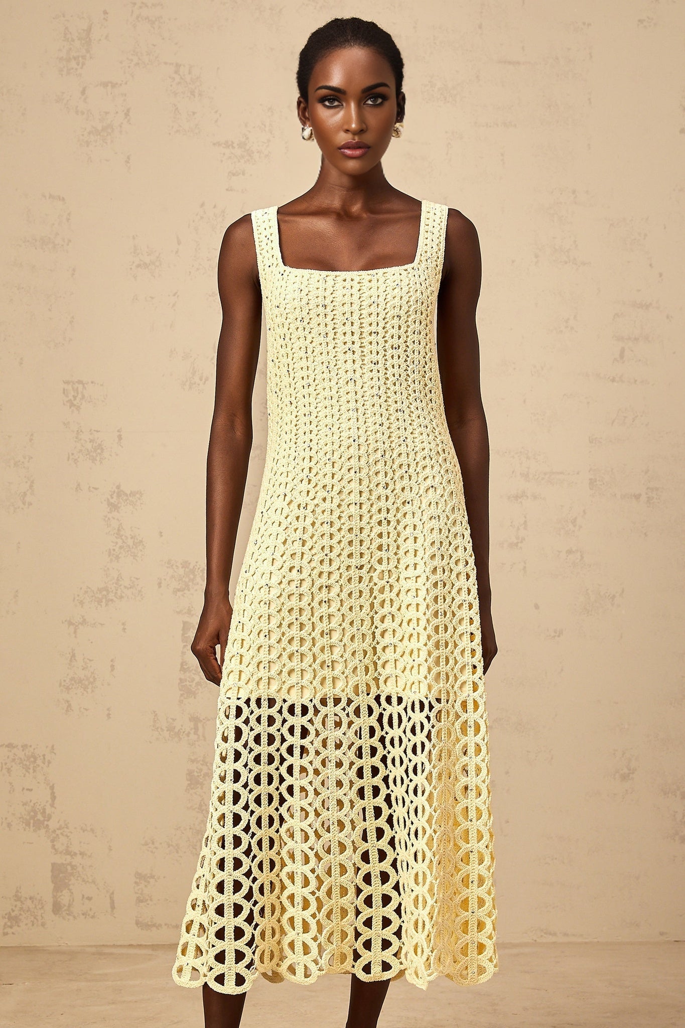 Woman wearing light yellow crochet knit midi dress with embellishments and cutout details standing against plain background