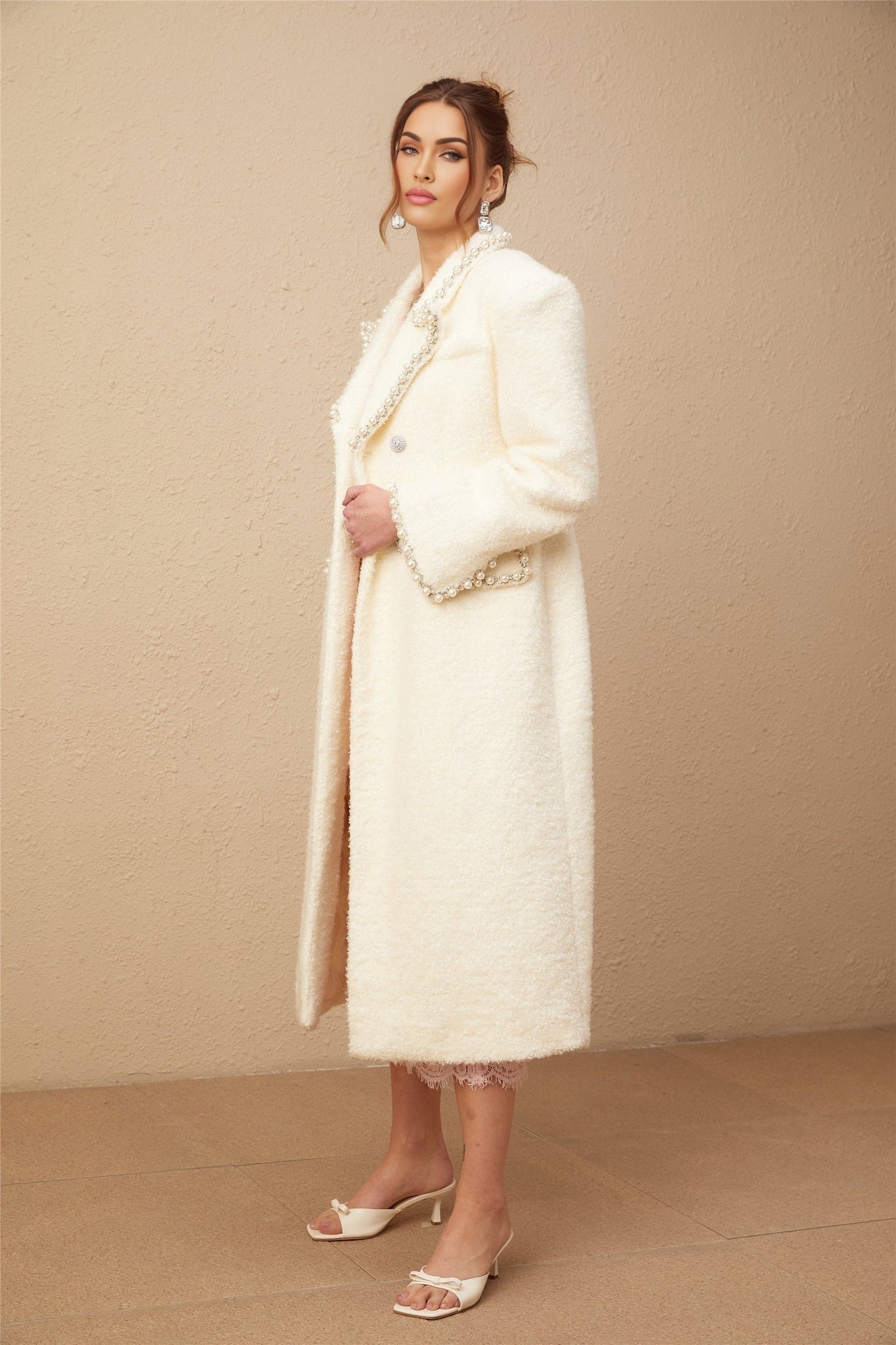 Woman wearing long beige tweed coat with faux pearl embellishments on collar and cuffs standing against plain background