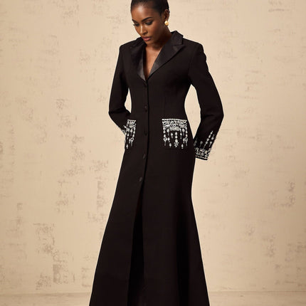 Woman wearing long black blazer dress with embellishments standing against plain background