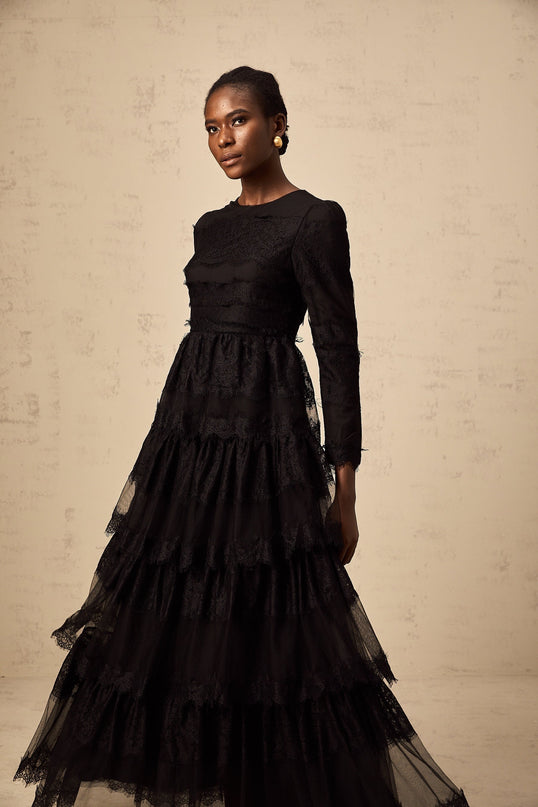 Woman wearing long black lace dress with tiered skirt and sheer tulle details standing against plain background