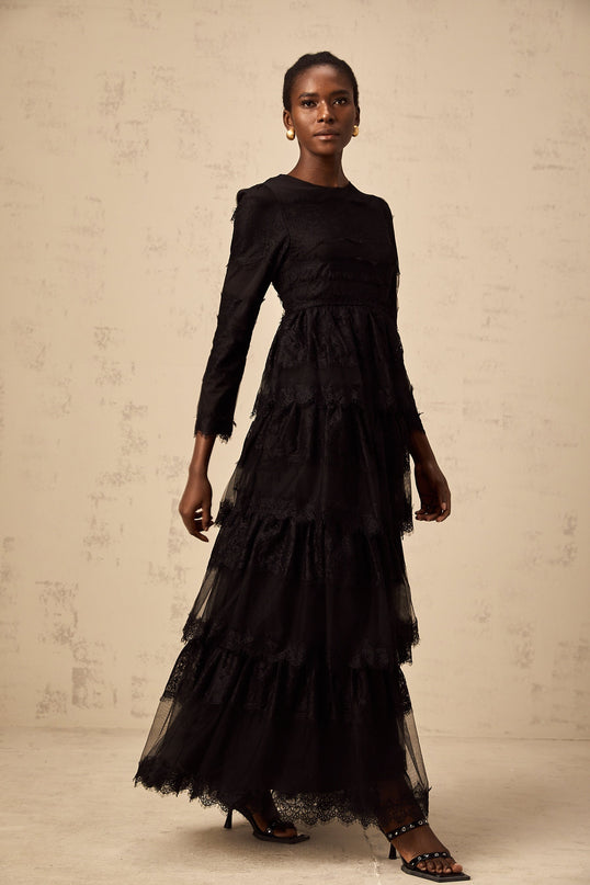 Woman wearing long black lace dress with tiered skirt and sheer tulle details standing against plain background